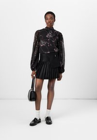 Blusa nera con motivo floreale e maniche a sbuffo, abbinata a una mini gonna plissettata nera. Gli accessori includono una borsa nera e mocassini con calzini bianchi.