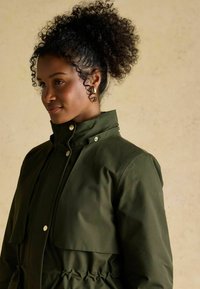 Oliwkowa wodoodporna kurtka z wysokim kołnierzem, zatrzaskami i ściągnięciem w talii, z dużą kieszenią z przodu. Teksturowane wykończenie.