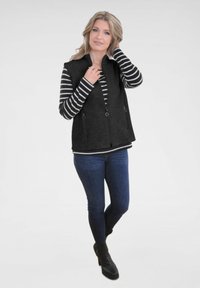 Zwarte ritsvest met een gestructureerd oppervlak, gedragen over een zwart-wit gestreept langslijfhemd, gecombineerd met donkere jeans en zwarte enkellaarsjes.