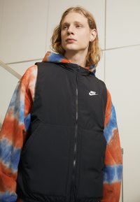 Crni puffer prsluk s kapuljačom, zatvaranjem na zip, i prednjim džepovima, nosi se preko tie-dye dukserice u narančastoj, plavoj i bijeloj boji. Nike logo vidljiv na prsima.