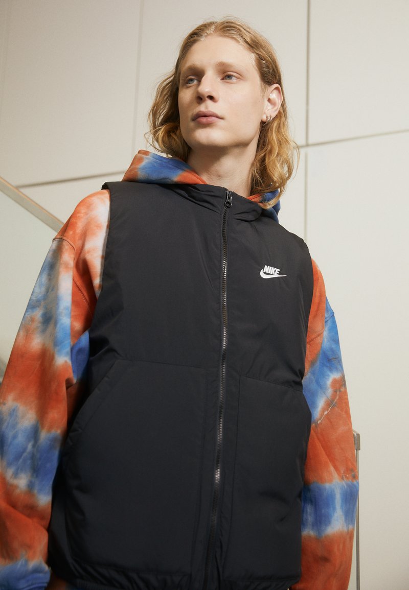 Crni puffer prsluk s kapuljačom, zatvaranjem na zip, i prednjim džepovima, nosi se preko tie-dye dukserice u narančastoj, plavoj i bijeloj boji. Nike logo vidljiv na prsima.