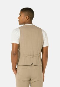 Beige op maat gemaakte gilet met een subtiele glans, minimale achternaad, verstelbare gespdetail op de taille, gedragen over een witte t-shirt.