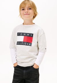 Grå T-shirt med korta ärmar och "TOMMY HILFIGER"-logotypen i svart och rött. Bärs över en vit långärmad tröja. Svarta jeans med fickor.
