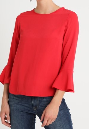 Mujer con una blusa roja brillante de manga larga con puños acampanados y jeans azules, de pie contra un fondo blanco simple.