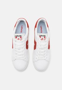Białe sneakersy z czerwonymi zamszowymi akcentami, z sznurowadłami, teksturowaną podeszwą i niebieską wkładką. Widoczne logo Levi's na języku.