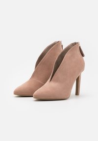 Botines de ante en rosa suave con punta afilada y un diseño de corte curvado. Presentan un cierre de cremallera en la parte posterior y un tacón alto y delgado.