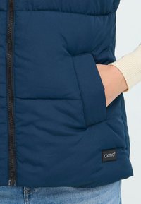 Tumši zila pūkurdžaka ar rāvējslēdzēju priekšā, plecu kabatu un logotipa etiķeti apakšmalā. Iezīmēta ar quilted tekstūru izolācijai.