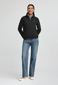 Sudadera negra con cremallera fabricada en material suave, combinada con jeans rectos azul claros y zapatillas beige. Diseño sencillo con un pequeño logo.