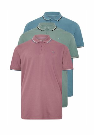 Mann mit muskulösem Körperbau, der ein grünes Poloshirt, schwarze Hose und weiße Sneakers trägt, steht vor einem einfarbigen Hintergrund.