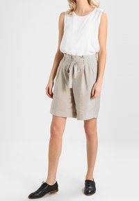 Top sin mangas blanco ligero combinado con pantalones cortos beige de cintura alta que cuentan con un cinturón atado. Unos zapatos negros con cordones completan el conjunto.