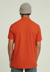 Polo shirt da uomo a maniche corte di un vivace colore arancione, realizzato in un tessuto morbido. Presenta un colletto classico e delicati polsini. Indossato con un berretto in maglia grigio chiaro.