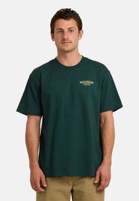 Camiseta de algodón verde con cuello redondo, mangas cortas y un pequeño logotipo gráfico en beige y blanco en la parte izquierda del pecho.