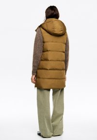 Mujer vistiendo un chaleco acolchado largo marrón sin mangas con capucha, un suéter marrón esponjoso, pantalones sueltos verde claro y zapatos negros, de pie con la espalda hacia el observador.