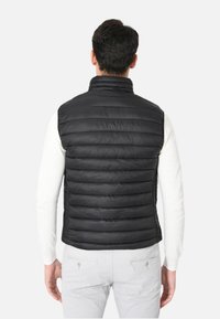 Gilet imbottito nero con colletto alto, design trapuntato e texture liscia. Indossato sopra una camicia bianca a maniche lunghe, che presenta una forma aderente.