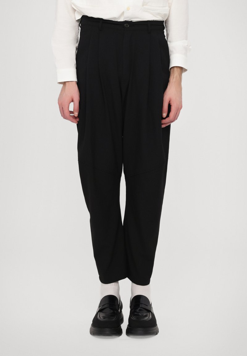 Pantaloni neri su misura con una vestibilità rilassata, dotati di vita alta, pieghe e una texture morbida. Abbinati a scarpe nere e calzini bianchi.