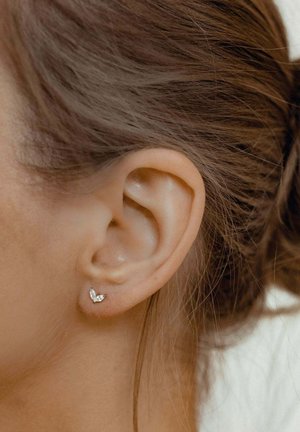 Pendientes de stud en forma de mariposa de plata, con tres pequeñas piedras preciosas transparentes, exhibidos en la oreja de una persona con cabello castaño.