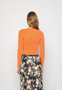 Tailliertes orangefarbenes Langarmoberteil mit rundem Halsausschnitt; kombiniert mit weit geschnittenen Hose im Blumenmuster auf dunklem Hintergrund.