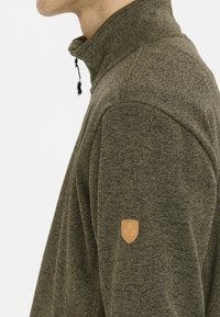 Grön fleecejacka med hög krage, strukturert tyg, dragkedjeförslutning och en beige läderlapp på vänster ärm.