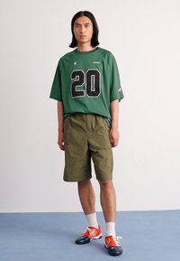 Young man wearing a green sports jersey with number 20, olive green shorts, white socks, and red and blue sneakers, standing on blue floor.