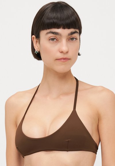 Top bikini triangolo marrone con spalline sottili. Tessuto liscio e design minimalista. Il modello ha i capelli corti e orecchini piccoli.