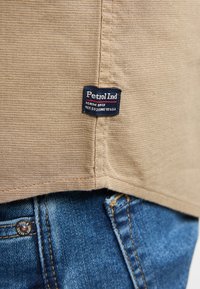 Beige katoenen stof met een gestructureerd oppervlak; bevat een zwart label met de tekst "Petrol Ind" dat in de naad is genaaid; onderliggende donkerblauwe denim is zichtbaar.