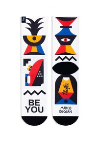 Sok in witte stof met kleurrijke geometrische patronen in rood, blauw, geel en zwart. De tekst luidt "WEES JIJ" en "MARCO OGGIAN." Zwarte teentoevoegsels.