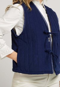 Gilet matelassé bleu marine avec une finition texturée, comportant une bordure en crochet le long de l'encolure, une fermeture à l'avant nouée et des poches latérales.