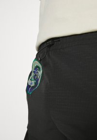 Sorte cargo-bukser med en tekstureret overflade, der har et farverigt broderet patch og justerbar snor i taljen.