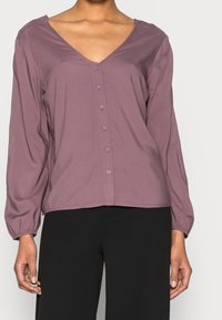 Mujer vistiendo una blusa de pico abotonada en malva con mangas largas y pantalones negros, de pie contra un fondo claro y liso.