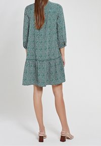 Zelené šaty se vzorem a vrstvenou sukní, s rukávy po tři čtvrtě a volným střihem. Nosí se s béžovými sandály na blokovém podpatku.