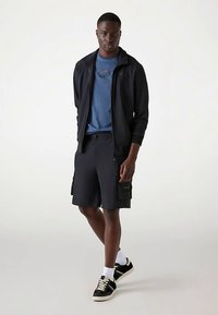 Must tõmblukk-jakk, sinine T-särk, tumedad cargo-lühikesed püksid küljetaskuga ning mustad spordijalatsid valgete tallade ja paeltega, kantud valgete sokkidega.