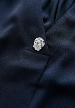 Tkanka w kolorze granatowym z diagonalną prążkowaną teksturą, ozdobiona błyszczącą, srebrną szpilką o zaokrąglonym kształcie z dokładnymi zdobieniami.