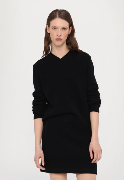 Maglione nero a maglia con scollatura a V e maniche lunghe, indossato sopra una gonna nera coordinata. Texture liscia e silhouette slanciata.