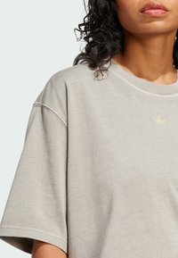 Vaaleanharmaa ylivertainen t-paita pyöreällä pääntiellä ja lyhyillä hihoilla, jossa pienikokoinen kulta-logo vasemman rinnan edessä.