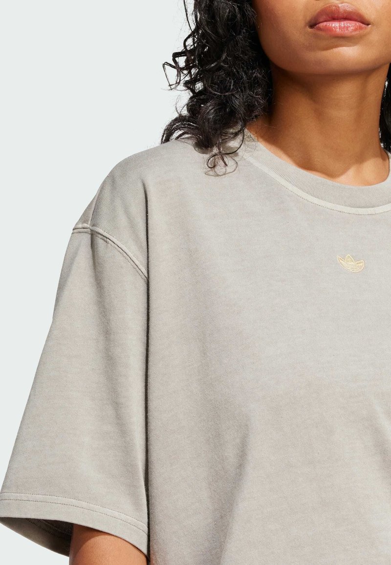 Camiseta oversize de color gris claro con cuello redondo y mangas cortas, con un pequeño logo dorado en la parte frontal izquierda del pecho.