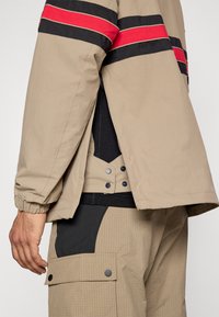 Ταν εξωτερικό μπουφάν με κόκκινες και μαύρες ρίγες, με ελαστικές μανσέτες. Από κάτω, μια μαύρη μπλούζα με κουμπιά και υφασμάτινα παντελόνια χακί με υφή.