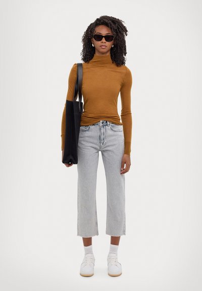 Brauner Rollkragenpullover, hellblaue cropped Jeans mit fransigem Saum, weiße Sneaker und eine schwarze Tote-Tasche. Model mit lockigem Haar und Sonnenbrille.