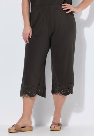 Vrouw die zwarte cropped broek draagt met bloemenlaser uitgesneden geschulpte zoom en bruine sleehakken, staand tegen een effen achtergrond.