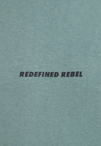 Licht teal katoenen t-shirt met de tekst "REDEFINED REBEL" in vetgedrukte zwarte lettertype, centraal gepositioneerd tegen een textuur achtergrond.