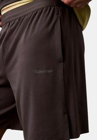 Pantalones cortos deportivos marrón oscuro con cinturilla elástica, que presentan un sutil logotipo de Calvin Klein en el lateral y una textura suave y ligera.