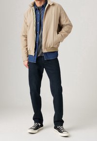 Man i beige bomberjacka över blå denimskjorta, grå t-shirt, mörka jeans och svarta canvas sneakers som står med handen i fickan.