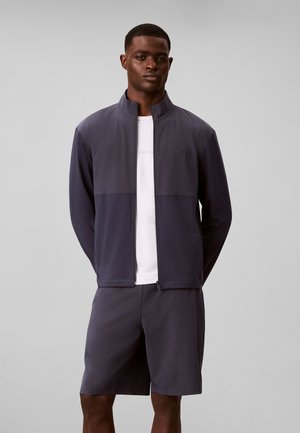 Un homme debout portant une veste zippée gris foncé, un short assorti et un t-shirt blanc, sur un fond uni gris clair.
