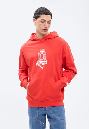 Jaunas vyras žiūri žemyn, dėvi raudoną megztinį su kapitonium ir „Österreich“ užrašu bei Puma logotipu, rankos kišenėje, dėvi mėlynas džinsines kelnes.