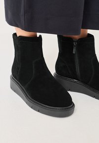 Czarne zamszowe botki za kostkę z niską platformą, subtelnym przeszyciem i bocznym zamkiem błyskawicznym. Gładka tekstura i czyste linie.