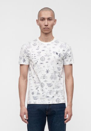 Homme portant un T-shirt blanc avec des croquis nautiques bleu marine et un jean bleu foncé, debout devant un fond uni.
