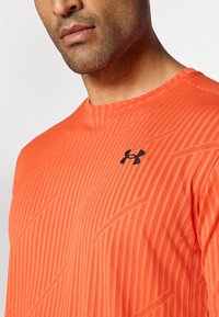Camiseta deportiva naranja con un sutil patrón texturizado y mangas cortas, que presenta un logo negro en el pecho y un escote redondo.