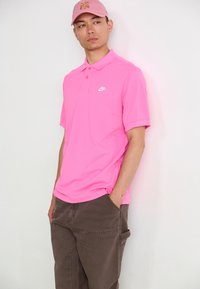 Rosa Nike Poloshirt mit einem Kragen mit zwei Knöpfen und kleinem Logo. Das Model trägt braune Cargo-Hosen, die eine entspannte Passform zeigen.