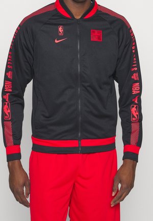 Mann trägt eine schwarze und rote Chicago Bulls Jacke mit Reißverschluss und NBA-Logos an Ärmeln und Brust, kombiniert mit roten Hosen.
