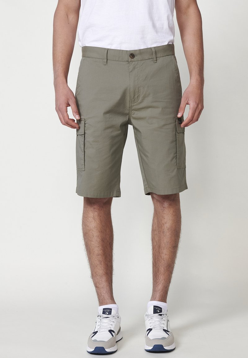 Uomo che indossa pantaloni cargo olive e sneakers bianche, in piedi contro uno sfondo chiaro e uniforme.