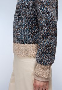 Wełniany sweter w ciemnym niebieskim, brązowym i kremowym kolorze, z teksturowanym wzorem, ściągaczami u dołu i na rękawach. Noszony na jasnych spodniach.